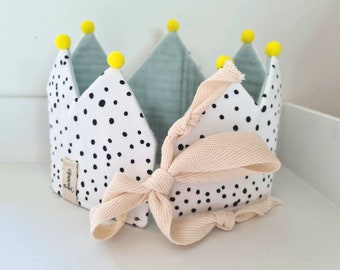 May include: A soft fabric crown with a white base and black polka dots. The crown has light blue interior panels and is topped with yellow pom-poms. A cream-colored ribbon is tied in a bow at the front. A small tag with text is sewn on the side.