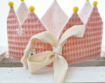 May include: A pink waffle-textured crown with yellow pom-poms and a white ribbon tied in a bow. The crown has a white daisy embroidered on the front.