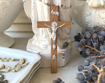 May include: A wooden crucifix with a silver Jesus figure, a small banner, and a decorative base. The cross is set against a backdrop of white decorative items and a bunch of dark blue berries.