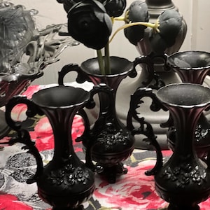 May include: A set of three small black vases with ornate handles and embossed floral detailing on the base. Each vase features a flared rim and a pedestal foot. One vase contains a cluster of artificial black flowers with yellow stems.