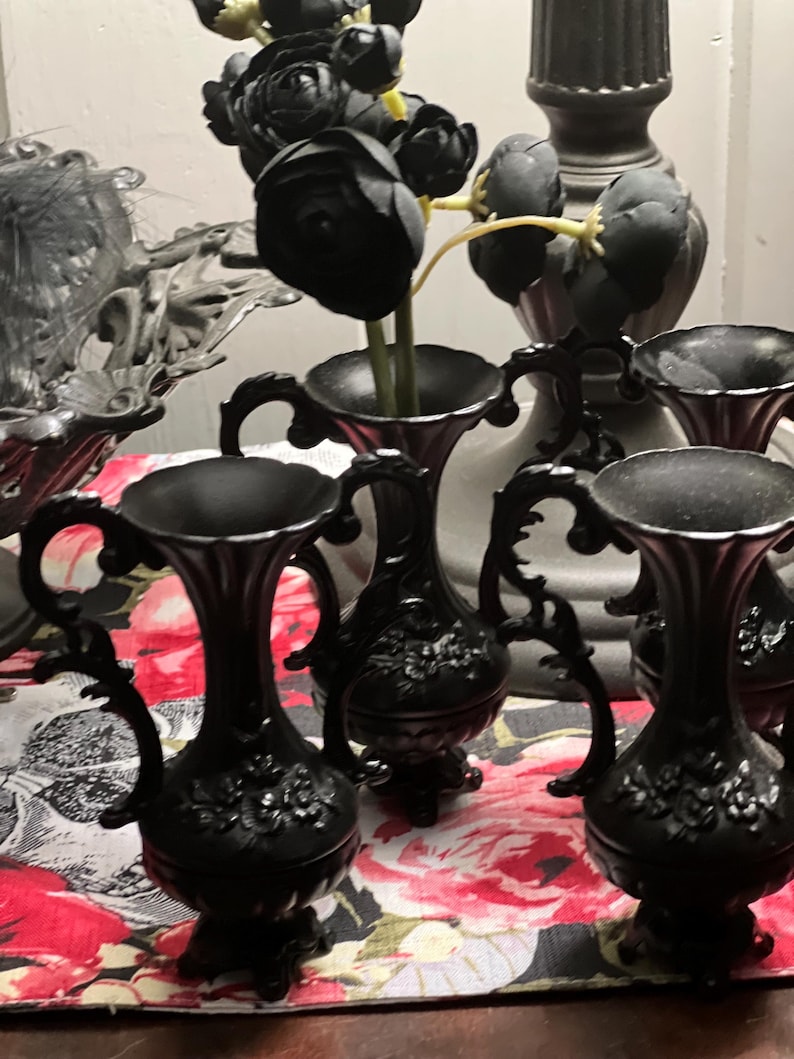 May include: A set of three small black vases with ornate handles and embossed floral detailing on the base. Each vase features a flared rim and a pedestal foot. One vase contains a cluster of artificial black flowers with yellow stems.
