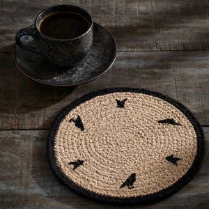 May include: A round, beige and black coaster with a crow design. The coaster has a black border and features six black crow silhouettes. A black coffee cup and saucer sit on a wooden table.