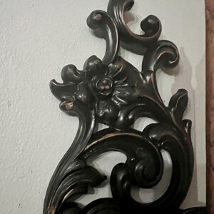 Pode incluir: Decora&ccedil;&atilde;o de parede ornamentada, castanho escuro, com um design floral e de rolagem. A pe&ccedil;a decorativa apresenta uma flor central e folhas estilizadas. As bordas t&ecirc;m um sutil sotaque dourado. Montado em uma parede branca.