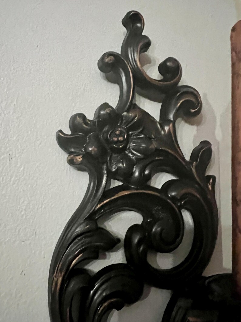 Pode incluir: Decora&ccedil;&atilde;o de parede ornamentada, castanho escuro, com um design floral e de rolagem. A pe&ccedil;a decorativa apresenta uma flor central e folhas estilizadas. As bordas t&ecirc;m um sutil sotaque dourado. Montado em uma parede branca.