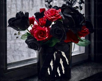 May include: A dark, gothic-themed floral arrangement in a black vase. The bouquet features vibrant red roses and deep black roses, accented with black foliage. A decorative skeleton hand detail adorns the vase, tied with a black ribbon. The arrangement sits on a dark windowsill.