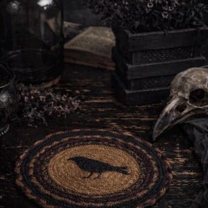 May include: A circular jute coaster featuring a black crow design. The coaster has a brown central area, surrounded by black and burgundy rings. The background includes a skull, dark bottles, and a wooden box.