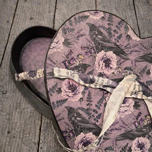 May include: Heart-shaped box with a floral and raven design. The box is open, revealing a black interior. The lid features a pattern of purple flowers, dark green foliage, and black ravens. A cream-colored ribbon is tied around the box.