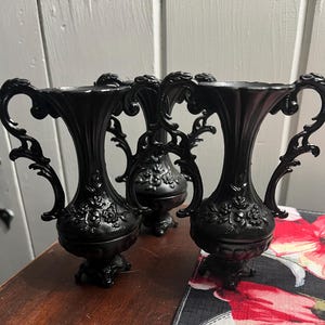 May include: Three matte black vases featuring ornate, scrolled handles and a raised floral relief pattern on the bulbous body. Each vase has a flared rim, a pedestal base, and a consistent dark finish, standing upright with symmetrical side handles.