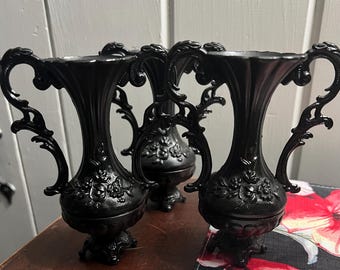 May include: Three matte black vases featuring ornate, scrolled handles and a raised floral relief pattern on the bulbous body. Each vase has a flared rim, a pedestal base, and a consistent dark finish, standing upright with symmetrical side handles.