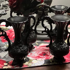 May include: A set of three matte black vases featuring ornate, scrolled handles and embossed floral detailing on the bulbous base. Each vase has a flared rim and stands on a small pedestal base, showcasing a consistent dark finish throughout.