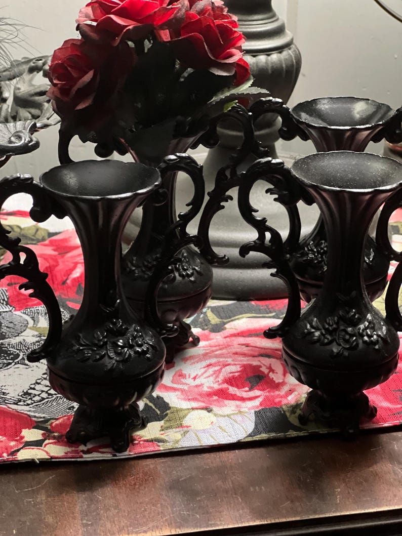 May include: A set of three matte black vases featuring ornate, scrolled handles and embossed floral detailing on the bulbous base. Each vase has a flared rim and stands on a small pedestal base, showcasing a consistent dark finish throughout.