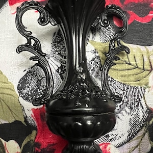 May include: A matte black decorative urn featuring a tall, fluted neck, two ornate scrolled handles on either side, and a bulbous base with embossed floral detailing. The vessel stands on a pedestal base with decorative feet and a scalloped rim.