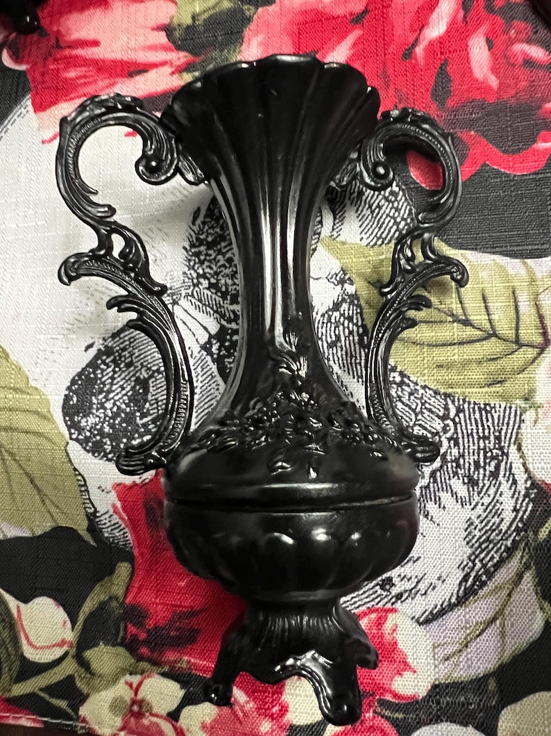 May include: A matte black decorative urn featuring a tall, fluted neck, two ornate scrolled handles on either side, and a bulbous base with embossed floral detailing. The vessel stands on a pedestal base with decorative feet and a scalloped rim.