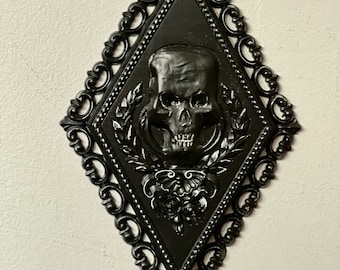 May include: A black diamond-shaped wall plaque featuring a central relief of a human skull surrounded by laurel leaf motifs and floral accents. The frame has an ornate, scrollwork border with a beaded edge and a small loop at the top for hanging.