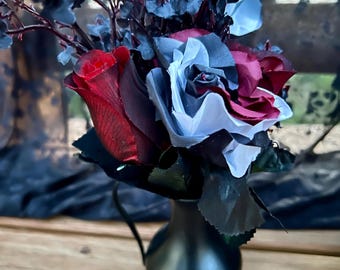 May include: A black vase displays a bouquet of artificial flowers. The arrangement features red, burgundy, and light blue roses, with dark blue foliage. The vase is matte black and has a handle. The flowers are arranged on a wooden surface.