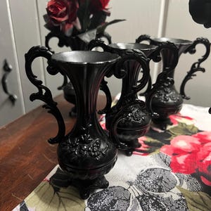 May include: A set of three small black vases with ornate handles and embossed floral detailing on the bulbous base. Each vase features a flared rim and stands on a pedestal base. The vases are arranged in a cluster, showcasing their matte finish.