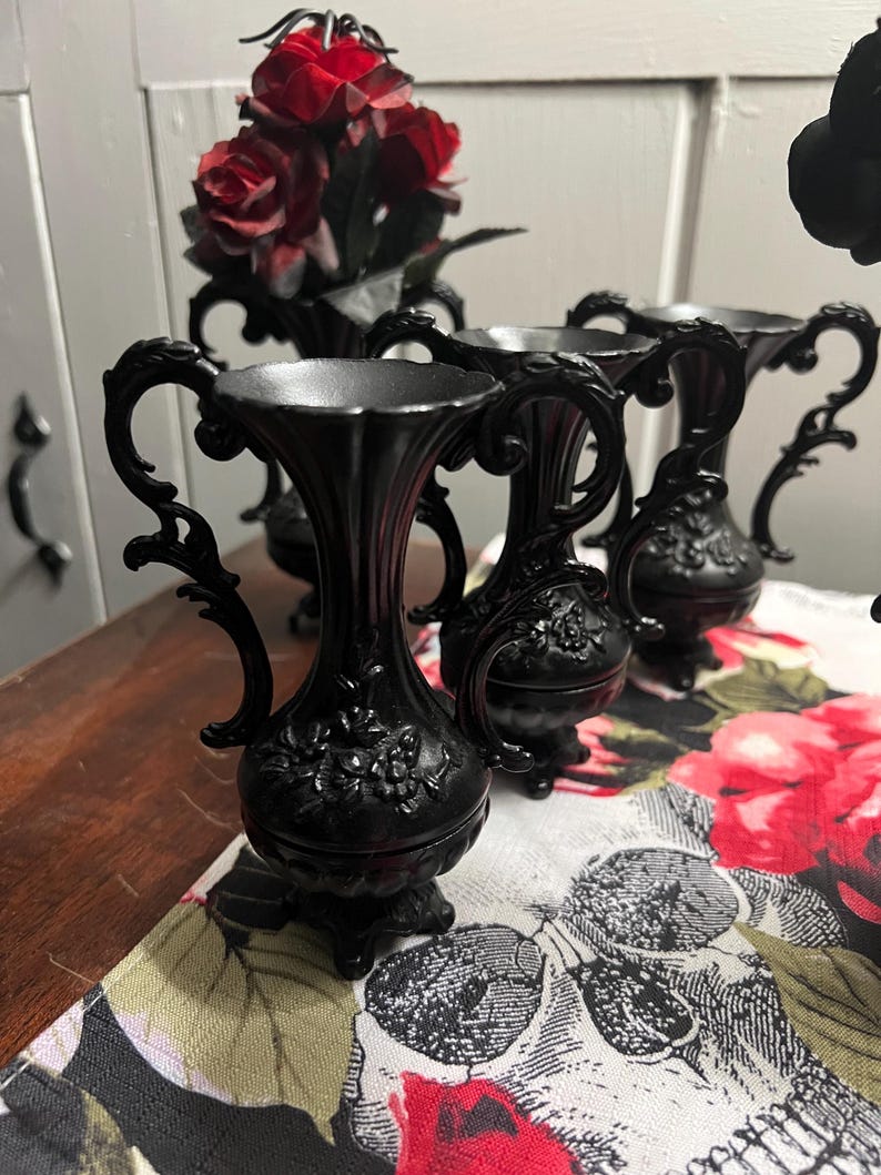 May include: A set of three small black vases with ornate handles and embossed floral detailing on the bulbous base. Each vase features a flared rim and stands on a pedestal base. The vases are arranged in a cluster, showcasing their matte finish.
