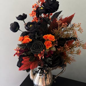 May include: A floral arrangement featuring black roses and orange blossoms in a silver-toned metal teapot with a handle, spout, and four decorative feet. The bouquet includes dark foliage, feathery orange plumes, and small orange berry-like clusters.