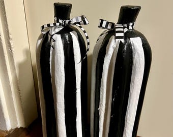 May include: Two decorative, tall, bottle-shaped objects. Each is painted with vertical black and white stripes. A black ribbon with a white stripe is tied around the neck of each bottle. The bottles are placed on a wooden surface.
