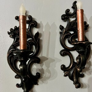 Pode incluir: Duas arandelas de parede ornamentadas, em bronze escuro, com casti&ccedil;ais cor de cobre e luzes de velas falsas. As arandelas t&ecirc;m um design detalhado e rodopiante e s&atilde;o montadas numa parede cinza claro. As luzes das velas t&ecirc;m um brilho suave e cintilante.