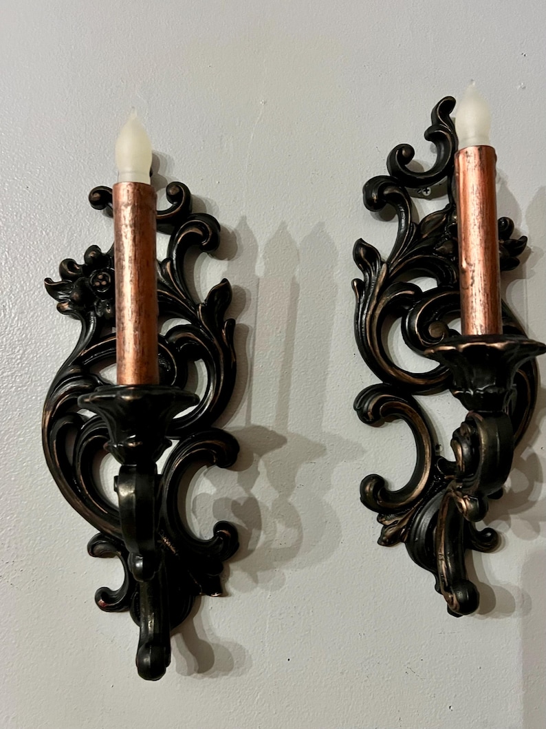 Pode incluir: Duas arandelas de parede ornamentadas, em bronze escuro, com casti&ccedil;ais cor de cobre e luzes de velas falsas. As arandelas t&ecirc;m um design detalhado e rodopiante e s&atilde;o montadas numa parede cinza claro. As luzes das velas t&ecirc;m um brilho suave e cintilante.