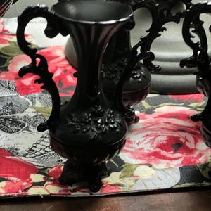 May include: A matte black vase featuring an ornate, flared rim and two curved, decorative handles on the sides. The body of the vase is adorned with a raised floral relief pattern and rests on a small, pedestal-style base with feet.