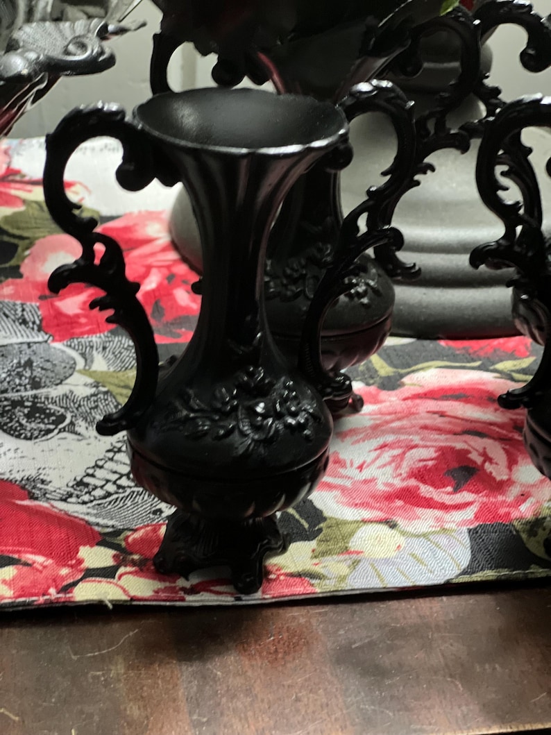 May include: A matte black vase featuring an ornate, flared rim and two curved, decorative handles on the sides. The body of the vase is adorned with a raised floral relief pattern and rests on a small, pedestal-style base with feet.