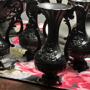 May include: A collection of matte black vases featuring ornate, raised floral relief patterns on the bulbous bodies. Each vase has a slender neck, a flared rim, and two curved handles on the sides, all supported by a small, decorative pedestal base.