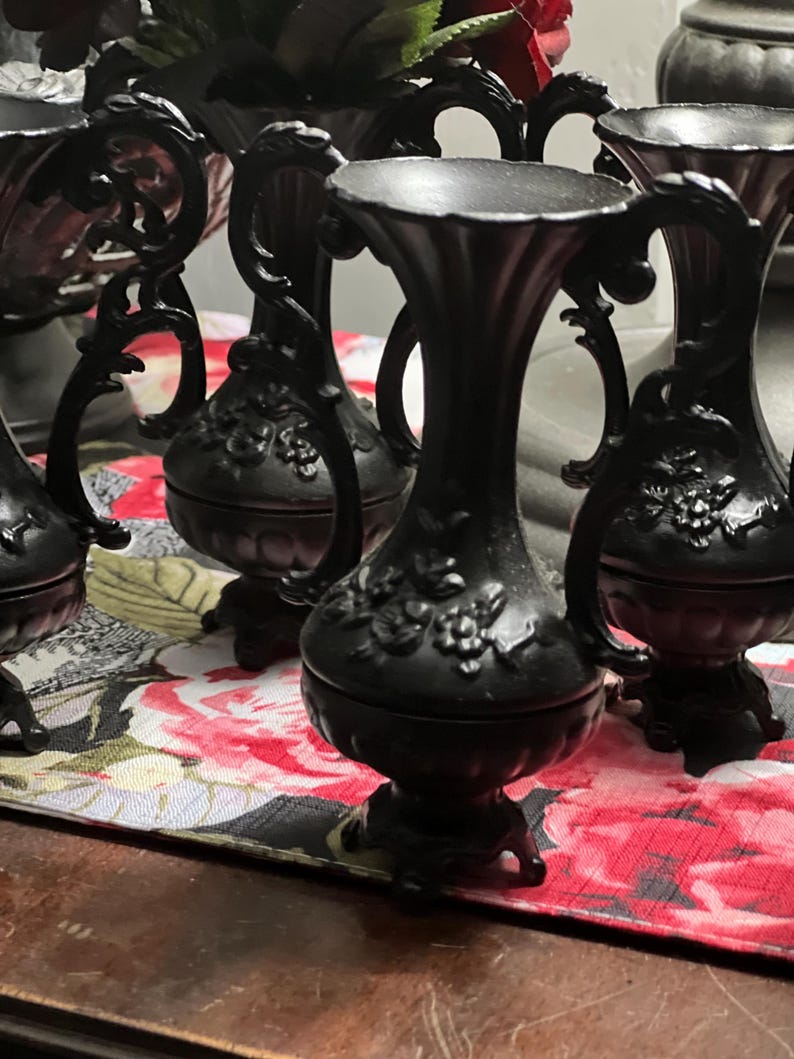May include: A collection of matte black vases featuring ornate, raised floral relief patterns on the bulbous bodies. Each vase has a slender neck, a flared rim, and two curved handles on the sides, all supported by a small, decorative pedestal base.