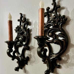 Pode incluir: Par de arandelas de parede ornamentadas com acabamento em bronze escuro. Cada arandela apresenta um design decorativo com volutas e cont&eacute;m uma vela falsa com base cor de cobre e ponta branca em forma de chama. As arandelas s&atilde;o montadas em uma parede branca.