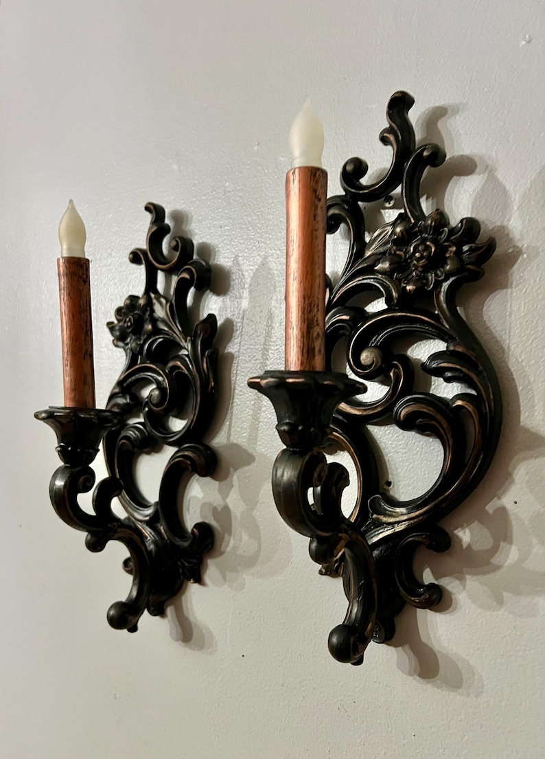Pode incluir: Par de arandelas de parede ornamentadas com acabamento em bronze escuro. Cada arandela apresenta um design decorativo com volutas e cont&eacute;m uma vela falsa com base cor de cobre e ponta branca em forma de chama. As arandelas s&atilde;o montadas em uma parede branca.