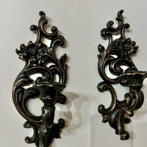 Pode incluir: Duas arandelas de parede ornamentadas em bronze escuro com detalhes florais e de volutas. Cada arandela apresenta um casti&ccedil;al e elementos decorativos, criando uma est&eacute;tica vintage. As arandelas est&atilde;o montadas numa parede cinza claro.