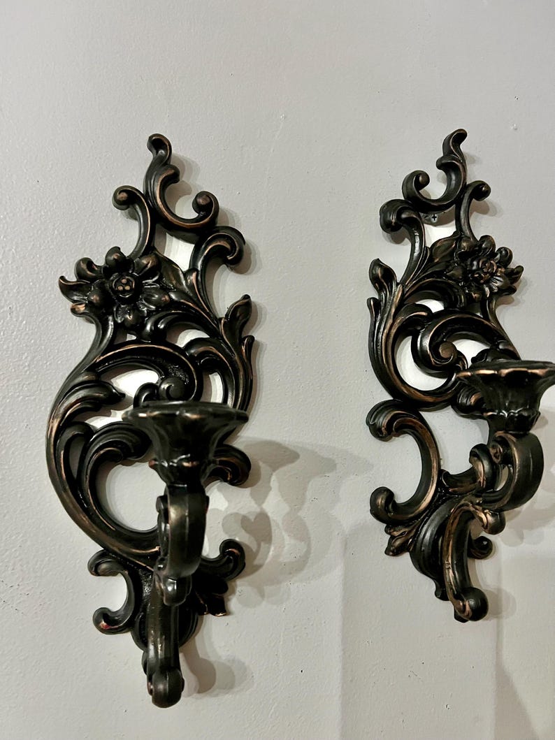Pode incluir: Duas arandelas de parede ornamentadas em bronze escuro com detalhes florais e de volutas. Cada arandela apresenta um casti&ccedil;al e elementos decorativos, criando uma est&eacute;tica vintage. As arandelas est&atilde;o montadas numa parede cinza claro.