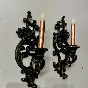 Pode incluir: Duas arandelas de parede ornamentadas, em bronze escuro, com um design floral e de rolagem. Cada arandela cont&eacute;m uma vela de cobre falso com uma chama LED tr&ecirc;mula, esbranqui&ccedil;ada. As arandelas s&atilde;o montadas em uma parede cinza claro.