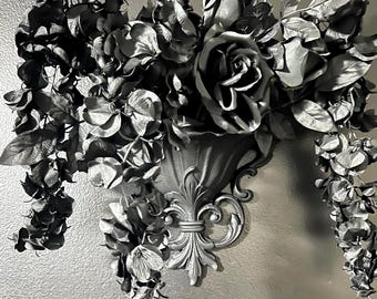 May include: A wall-mounted black planter featuring a decorative fleur-de-lis base, filled with an arrangement of dark black artificial roses and cascading floral foliage that hangs down from both sides of the central container.