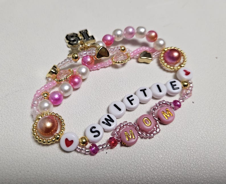 Pu&ograve; includere: Un braccialetto di perline rosa e bianche con le parole "SWIFTIE MOM" scritte in perline bianche. Il braccialetto ha anche ciondoli a forma di cuore dorati e un ciondolo dorato che dice "SL".