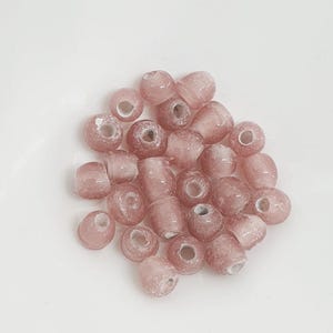 May include: A selection of pink glass beads in diverse shapes, including round, cylindrical, and irregular forms. The beads have a translucent quality, with visible internal textures and a slightly frosted appearance. They are scattered on a white surface.