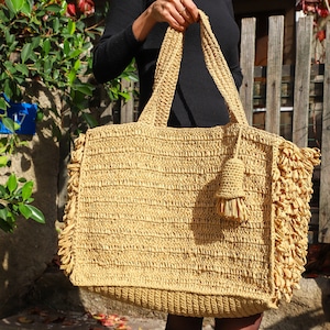 May include: A large, beige straw tote bag with a fringe trim and a tassel. The bag is made of woven straw and has a rectangular shape.