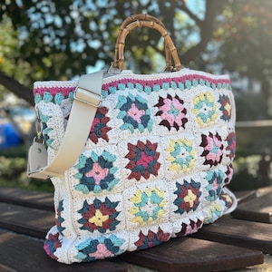 May include: A white crocheted tote bag with a colourful granny square pattern. The bag has a bamboo handle and a white canvas shoulder strap.