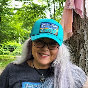 May include: Teal trucker hat with a mountain graphic and the text "Mountain Blaze". The hat is worn by a person with long gray hair, wearing a gray t-shirt with a matching mountain graphic.