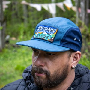 Peut inclure: Casquette bleue à cinq panneaux avec une visière plate. La casquette arbore un écusson rectangulaire avec l'inscription "Mountain Blaze" et une scène de montagne. Elle possède un panneau latéral en mesh et une fermeture à bouton-pression. La personne porte une veste noire.