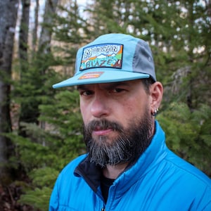 May include: A light blue and gray flat-brimmed cap with the text "Mountain Blaze" on a patch. The cap has a light blue brim and a gray mesh back. The person in the image is wearing a blue jacket.