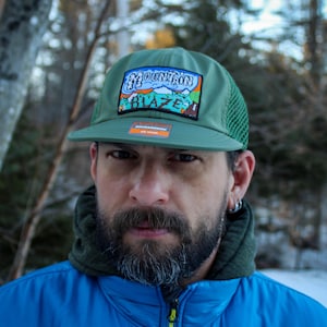 May include: A green trucker hat with a mesh back and a flat brim. The hat features a patch with the words "Mountain Blaze" and a mountain graphic. The hat is worn by a person in a blue jacket.