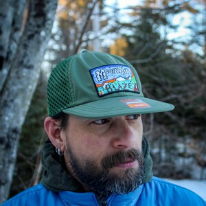 May include: Olive green trucker hat with a mesh back and a flat brim. The hat features a colorful patch with the text "Mountain Blaze" and a mountain graphic. The hat is worn by a person.