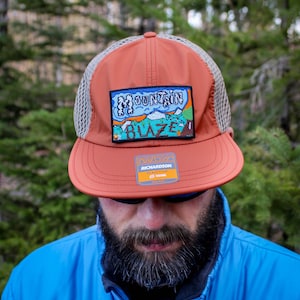 May include: A burnt orange and tan trucker hat with a rectangular patch that reads "Mountain Blaze" with a mountain scene. The hat has a curved brim and a mesh back. The brand tag is visible.