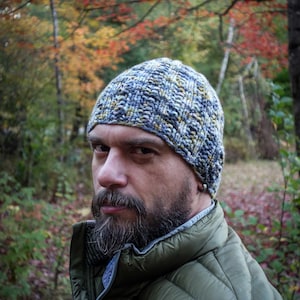 May include: A knitted beanie in shades of blue, yellow, and gray. The hat is worn by a person wearing a green quilted vest. The background features a forest with autumn foliage.