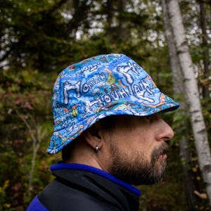 May include: A blue and orange bucket hat with a colorful, abstract design featuring the words "Mountain" and "Town" repeated. The hat is worn by a person with a beard.