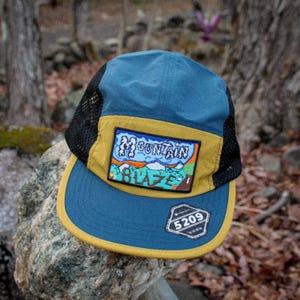 May include: A five-panel cap featuring a blue crown and brim with a mustard yellow front panel and black mesh side panels. A rectangular embroidered patch displays a mountain landscape with the text Mountain Blaze. A black sticker reads 5209.