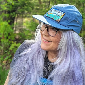 May include: A blue five-panel cap featuring a rectangular patch on the front with a graphic illustration of mountains, trees, and the text "MOUNTAIN HAZE" in a stylized font. The cap has a flat brim and a small metal eyelet on the side panel.