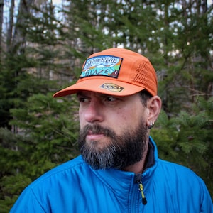 May include: Orange cap with a mesh side panel and a curved brim. The cap features a rectangular patch with the words "Mountain Blaze" and a mountain graphic. A small, circular logo is visible on the front.